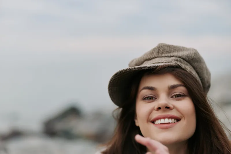 Smiling young woman with long brown hair wearing a stylish cap outdoors on a cloudy day. Close-up portrait with a blurred natural background. Concept of happiness and casual fashion.