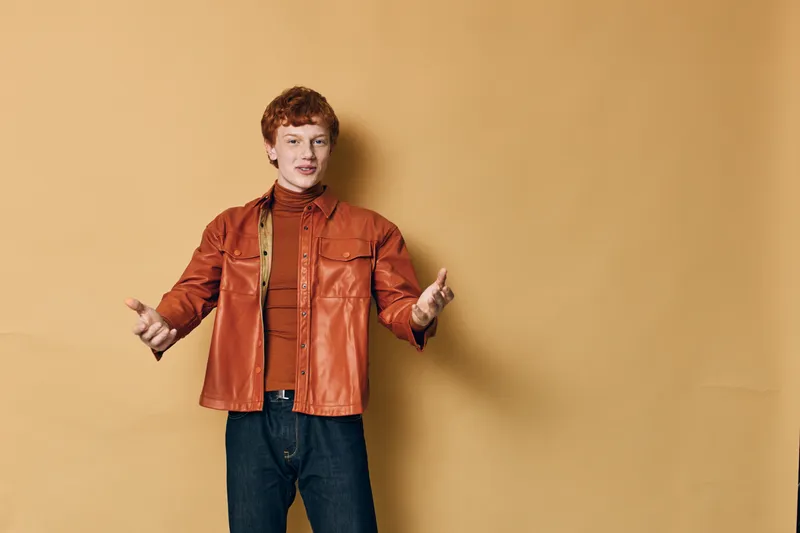 Young man in orange jacket and turtleneck standing with open arms against beige background for presentation or welcoming gesture and casual style.