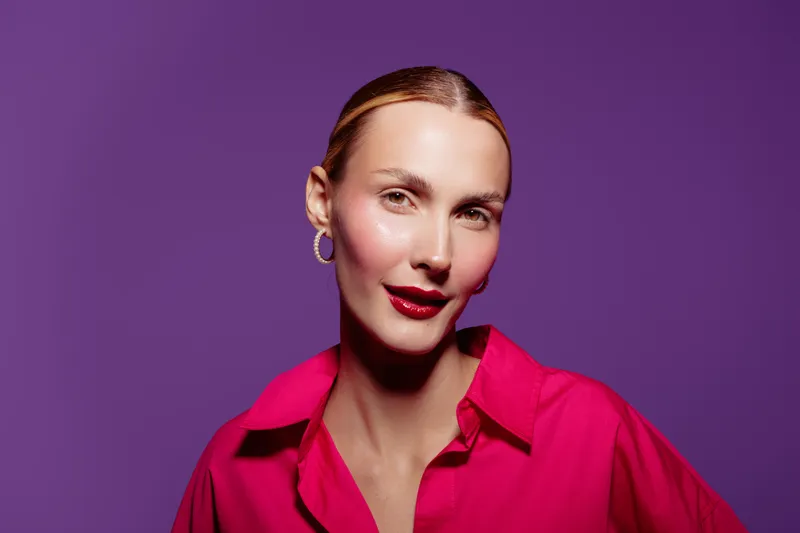 Portrait of confident woman with red lipstick and hoop earrings wearing bright pink shirt on purple background. Concept of fashion, beauty, and modern style with vibrant colors.