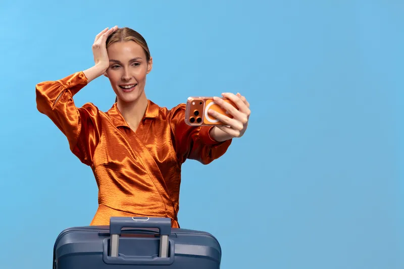 Young woman with suitcase takes selfie on smartphone against blue background. Travel concept with happy woman in orange blouse preparing for trip or vacation.