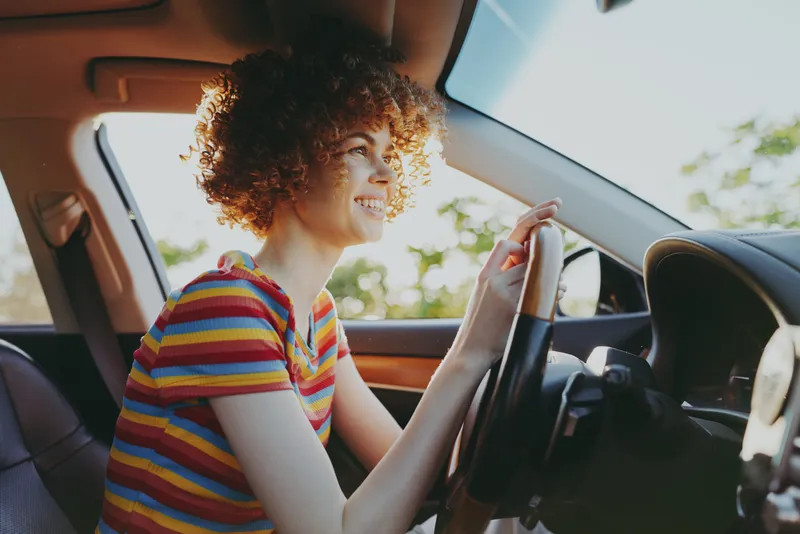 Woman driving a car in a casual lifestyle shot, wearing a striped shirt, warm retro film color, bright daylight, candid moment, independent traveler, everyday mobility