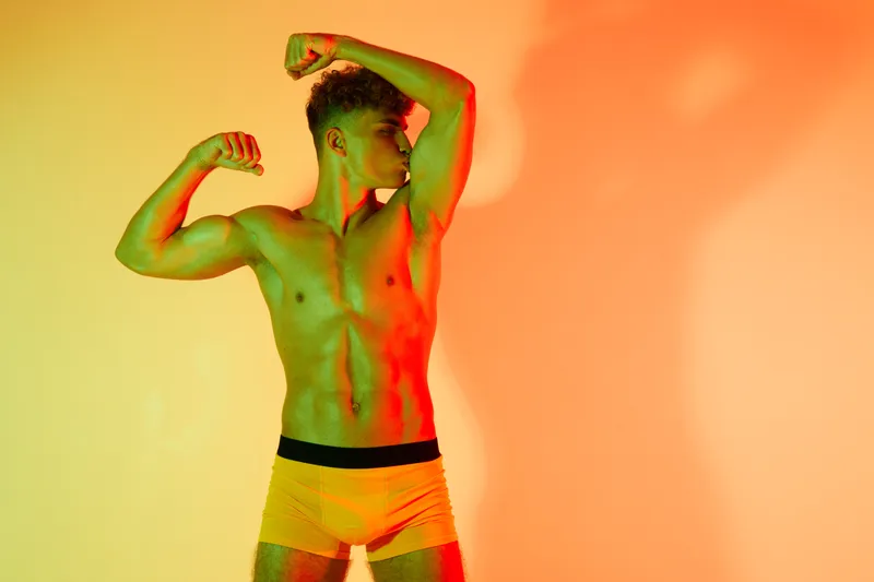 Athletic young man posing shirtless in yellow shorts flexing biceps with artistic contrast lighting for body fitness health and sport concept in studio background.