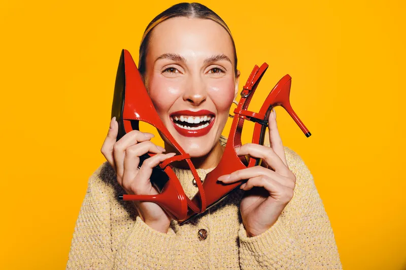 Happy woman with bright red lipstick holds red high heel shoes near face, smiling against vibrant yellow background. Concept of fashion, style, beauty, and joyful emotions.