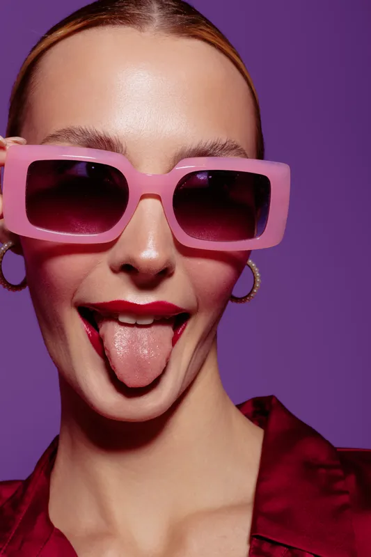 Young woman with pink sunglasses and red lipstick sticks out tongue on purple background. Playful expression, fashion style, and vibrant colors create fun mood.