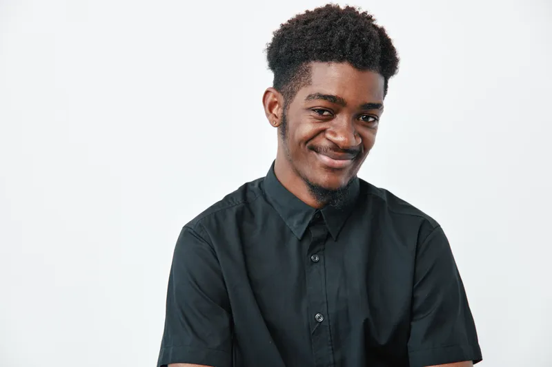 Young African American man with dark skin and curly hair wearing black shirt smiling confidently on white background. Portrait of happy male with casual style and positive expression.