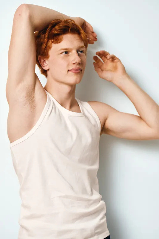 Young man with red hair and visible armpits wearing white tank top posing with arms raised against plain background for portrait or fashion concept.