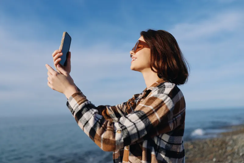 Woman holding a smartphone aloft by the seashore, capturing a moment with ocean breeze, wearing sunglasses and a plaid shirt, against a bright sky