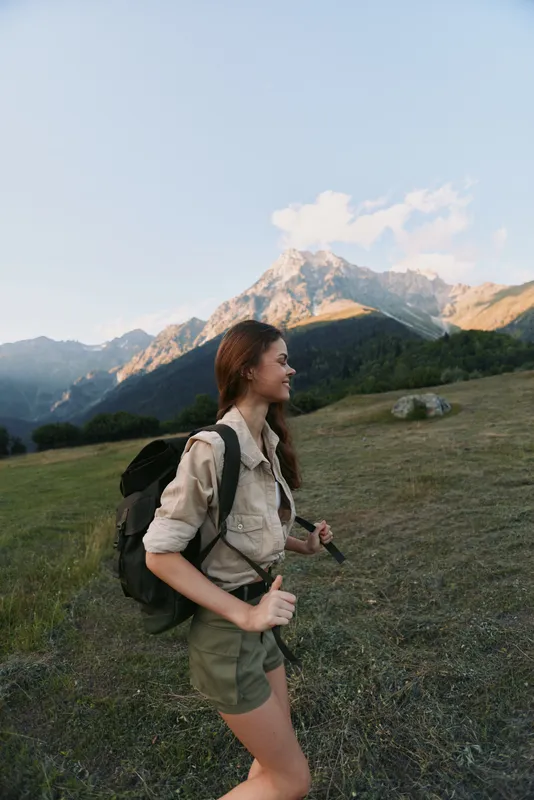 Woman hiker with backpack walks through alpine meadow, enjoying outdoor adventure, smiling, wearing practical clothes, majestic mountains in the background during daytime.