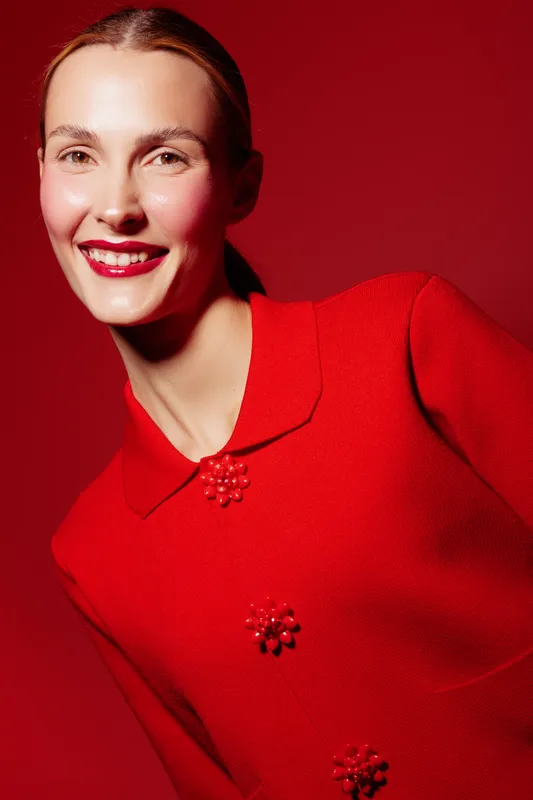 Smiling woman in red sweater poses against red background. Bright makeup and glossy lips highlight her joyful expression. Concept of fashion, beauty, and vibrant style.
