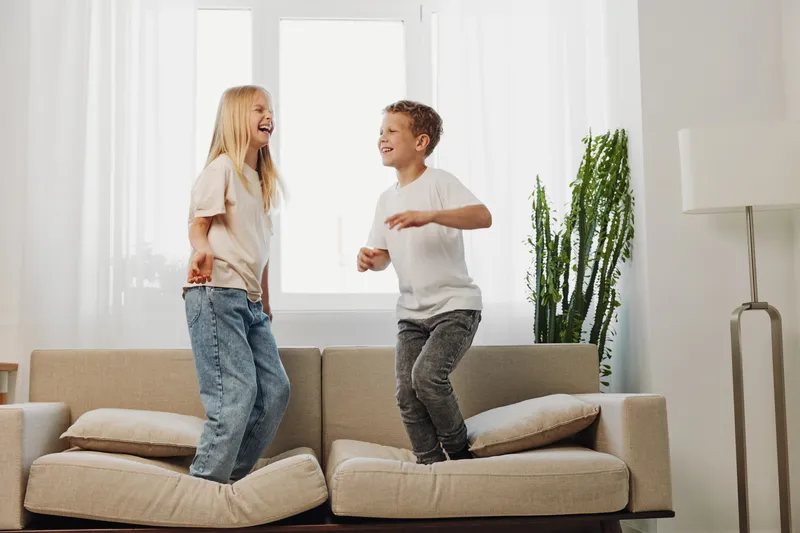 Happy children jumping on sofa in bright living room, laughing and enjoying playful moment together. Concept of childhood fun, energy, and family lifestyle indoors.