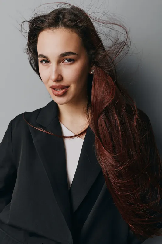 Woman with long hair and natural makeup wearing black blazer posing against gray background with confident expression and wind blowing through hair in studio portrait.