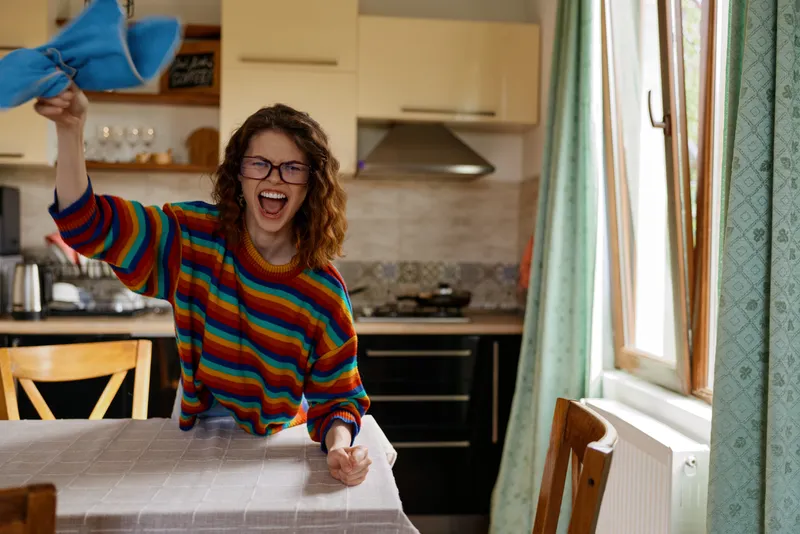 Cheerful woman with curly hair in a striped sweater laughs while tossing a blue cloth in a bright kitchen, standing by a counter. A candid lifestyle moment of home cleaning energy.