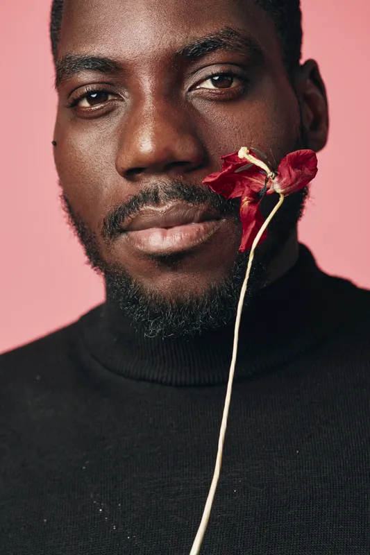 African man with dark skin and beard in black turtleneck holding dried red flower near face on pink background. Creative conceptual portrait with visual style and closeup expression.