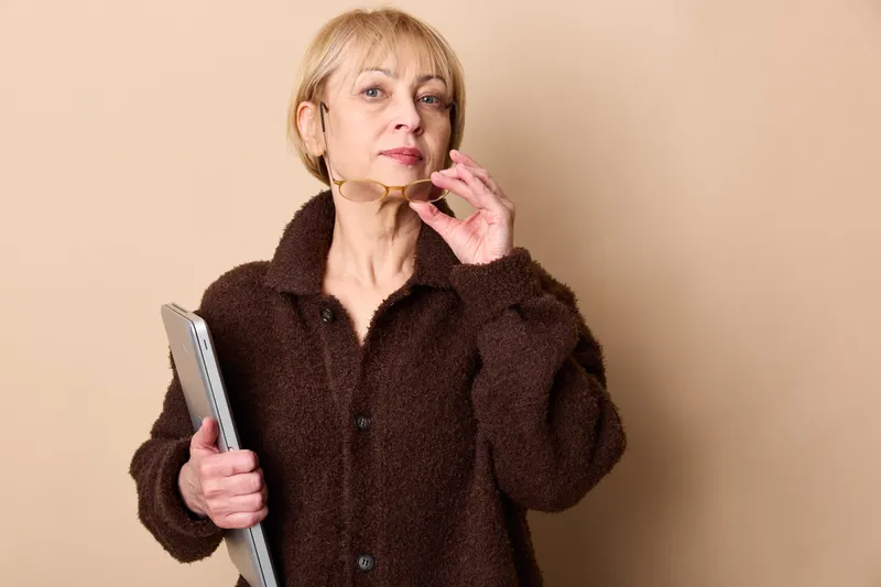 Mature woman applying white face cream with fingers, holding a phone in studio, soft lighting, beige background, close up portrait, skincare wellness and selfcare concept