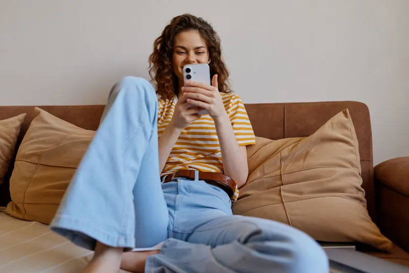 Casual young woman with curly hair sits on a cozy sofa at home, scrolling her phone and enjoying a relaxed moment in soft indoor light