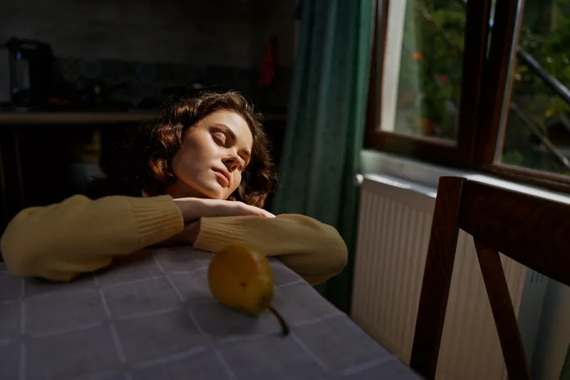 Calm woman rests her head on a table in a cozy home interior, soft window light highlights a contemplative mood, a quiet everyday moment indoors, solitary simple scene, warm tones