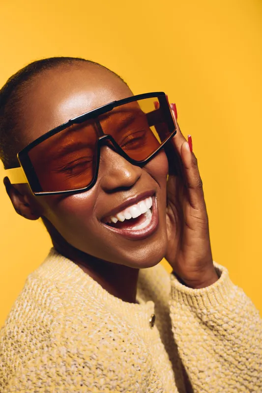 Happy African woman with dark skin wearing large sunglasses and a knitted sweater poses smiling on yellow background. Concept of fashion, beauty, joy, and stylish accessories.