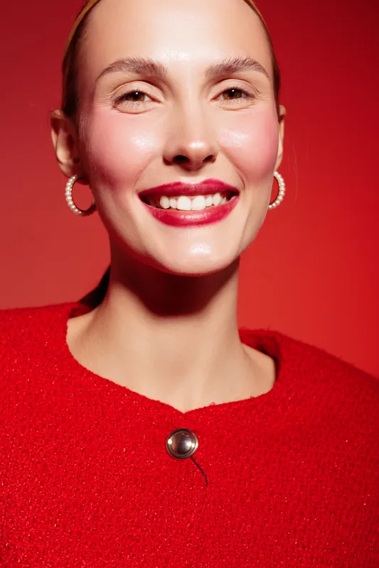 Smiling woman with bright red lipstick and earrings wears red sweater against red background. Close-up portrait with natural skin glow and joyful expression. Concept of beauty and fashion.