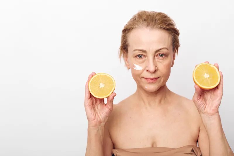 Mature woman with natural skin holds two halves of fresh orange in hands, with cream applied on her cheek against white background. Concept of skincare, vitamin c, and healthy lifestyle.