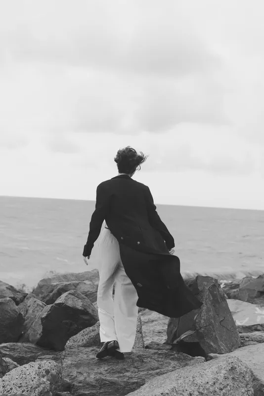 Silhouette of a figure standing on a rocky shoreline, wind swept coat flowing, sea in the background, monochrome scene conveying solitude, exploration and resilience by the coast, moody atmosphere