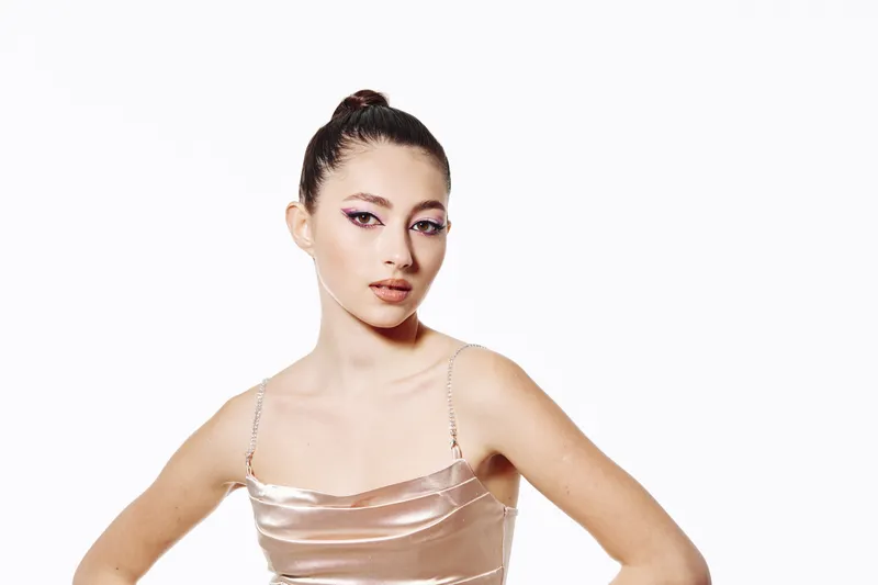 Fashionable young woman with sleek bun and shimmering makeup wearing a light champagne slip dress posing confidently on white background, beauty concept creative style.