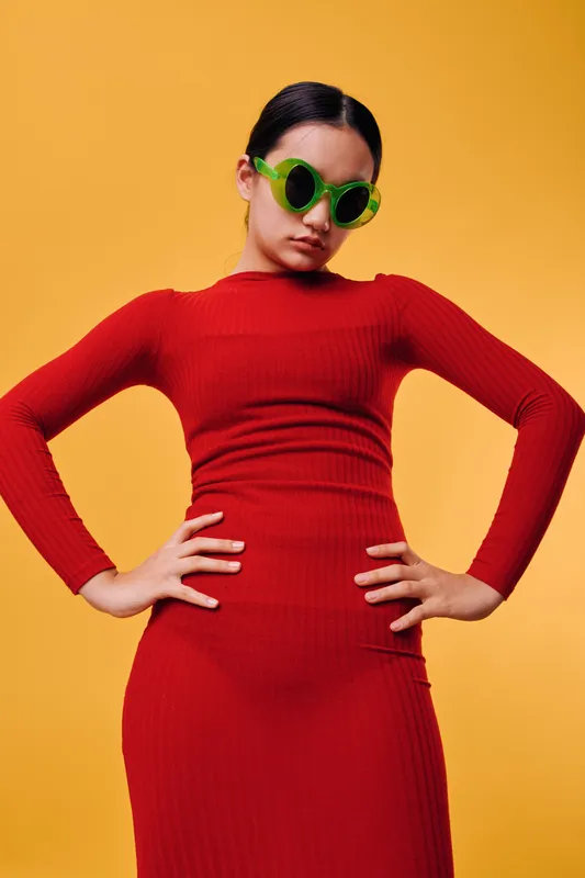Woman in red dress and green sunglasses posing with hands on hips against yellow background. Fashion model with sleek hairstyle and confident posture in studio photo shoot.