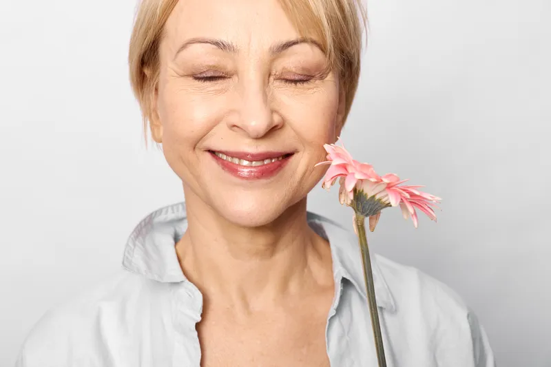 Woman smiling with eyes closed holding a pink flower in closeup portrait, calm expression, soft studio light, white background, gentle beauty, skincare care