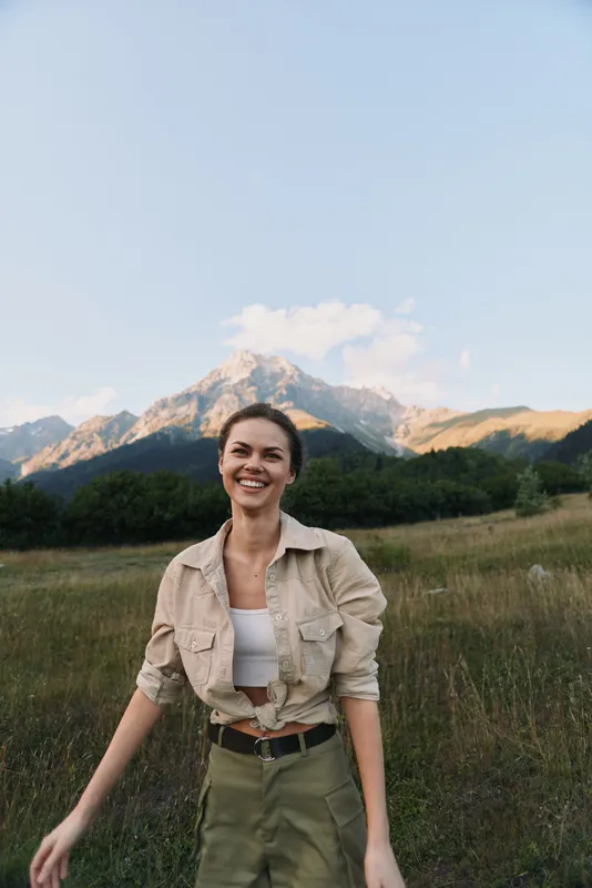Smiling woman outdoors standing in grassy field with mountains in background natural light casual outfit relaxed lifestyle happy expression