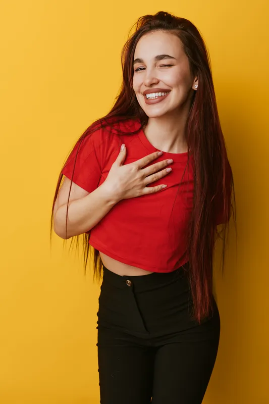 Young woman with long hair smiling and winking with hand on chest wearing red shirt and black pants standing against yellow background. Positive emotion and casual style portrait.