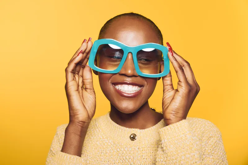 Smiling black woman with dark skin wearing oversized blue sunglasses and yellow sweater stands against bright yellow background. Concept of fashion, style, and joyful urban lifestyle.