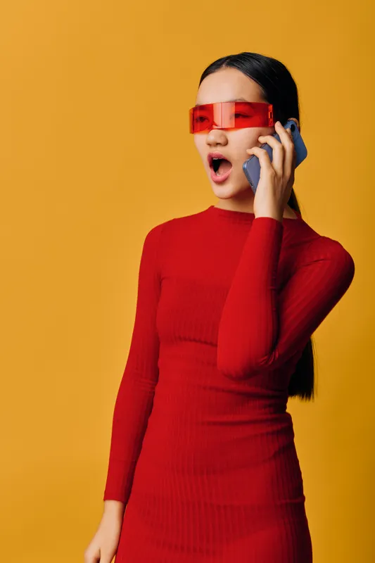 Young woman in red dress and futuristic sunglasses talking on phone with surprised expression on yellow background. Modern style, communication, technology and fashion portrait.