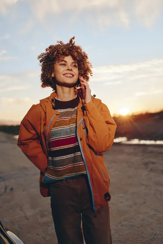 Woman lifestyle outdoors at sunset, curly hair, smiling, wearing jacket, she stands on rural road, warm golden light, gentle breeze, distant mountains softly blurred, relaxed stand