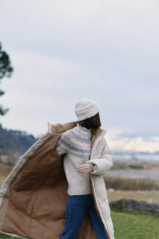 Woman outdoors wearing a warm coat, knit sweater and beanie, wind catching her coat as she stands in an open landscape with cloudy sky and distant fields