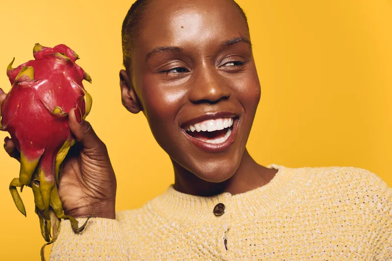 Smiling African woman with dark skin holds fresh dragon fruit against yellow background. Concept of healthy lifestyle, natural food, nutrition, and vibrant positive mood in studio lighting.