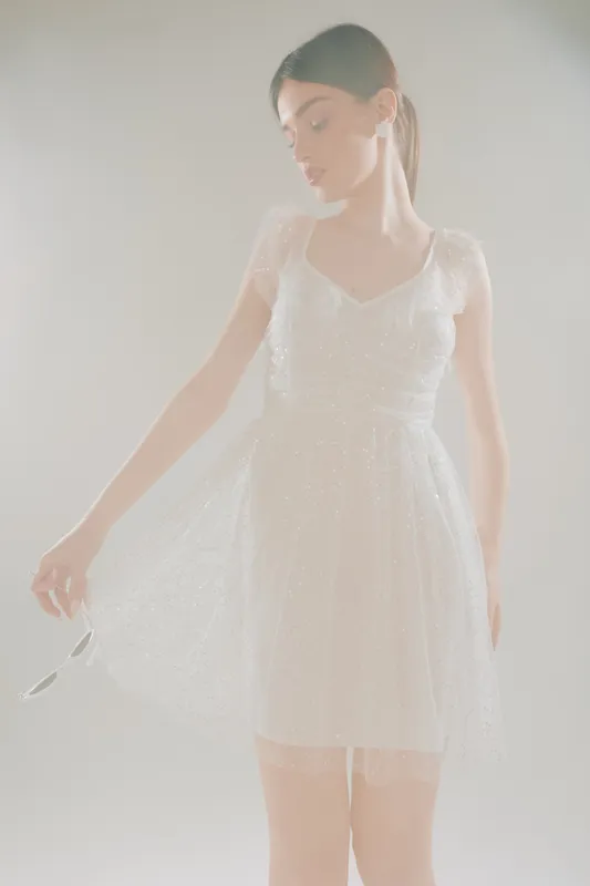 Young woman in a white lace dress poses gracefully holding sunglasses. Soft lighting highlights her beauty and fashion style on a neutral background. Concept for elegance and style.