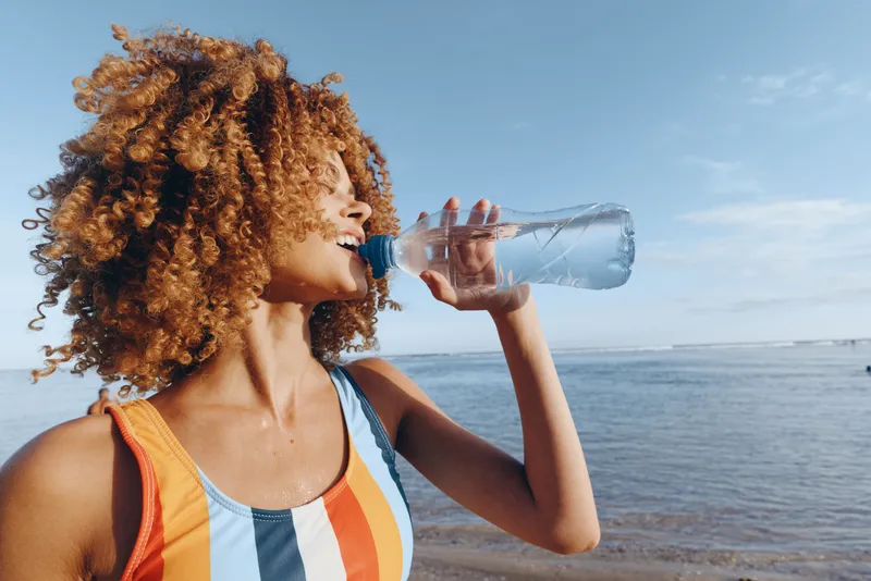Curly haired woman drinking water from a plastic bottle outdoors by the ocean, summer beach breeze, soft sunlight, relaxed profile, blue sky and calm sea background in clear weather