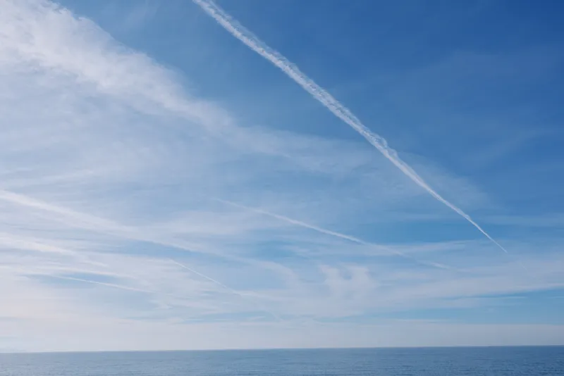 Clear blue sky above a calm sea, distant horizon, and white contrails crossing the expanse, creating a serene coastal atmosphere.