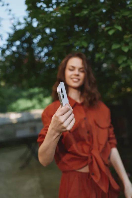 Cheerful woman outdoors, smiling while extending a phone for a selfie, sunlit moment, relaxed pose, warm tones, leafy background, lifestyle mood, candid portrait