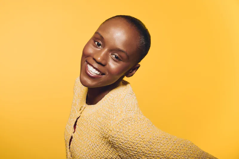 Smiling black woman with dark skin wearing a yellow sweater poses against a yellow background. Studio portrait with joyful expression and warm lighting for lifestyle concept.