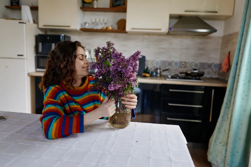 Smiling woman in a colorful striped sweater holds a bouquet of purple flowers at a kitchen table. Cozy home setting captures everyday life and kitchen mood.