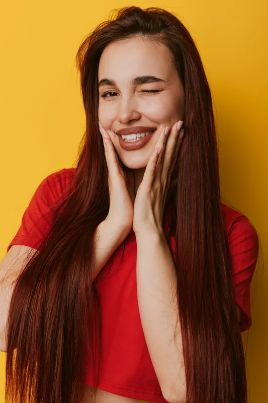 Young woman with long hair wearing red shirt winking and smiling with hands on face against yellow background. Portrait, beauty, happiness, casual style, confident female model.