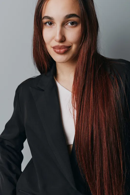 Young woman with long straight hair wearing black blazer and white top posing with confident smile against gray background in studio portrait for fashion or business concept.