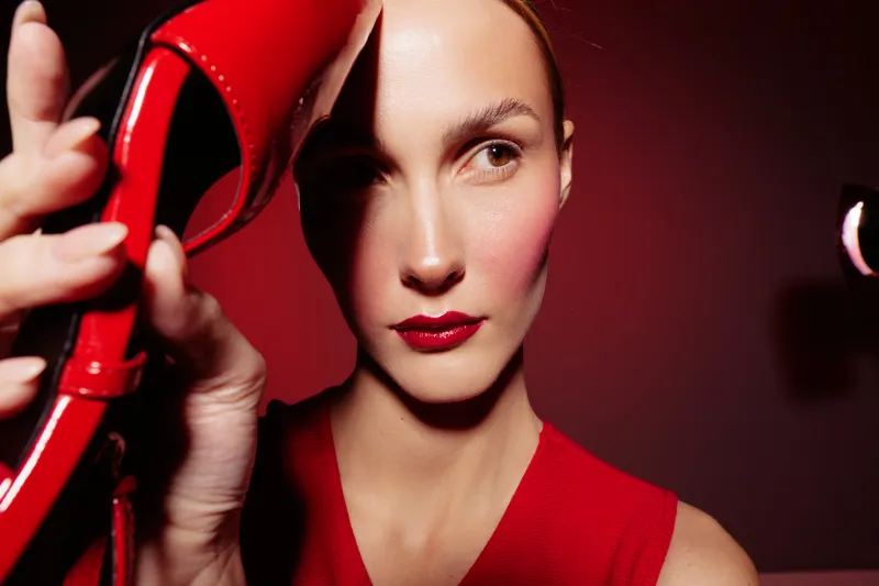 Close-up of a woman with red lipstick and red dress holding a red high heel shoe near her face. Dramatic lighting creates strong shadows on a red background. Concept of fashion and style.