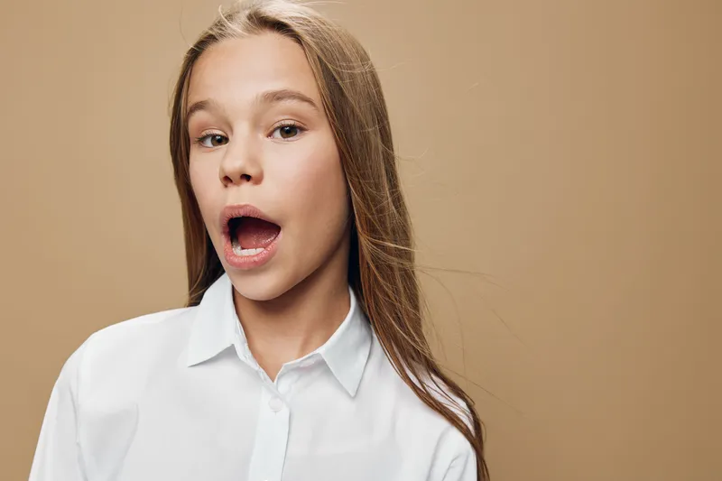 Girl with long hair and open mouth wearing white shirt on beige background. Portrait of surprised child with expression and casual clothing for emotion and communication.