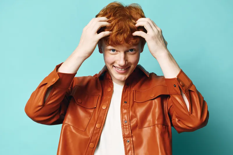 Redhead young man with curly hair smiling and running fingers through hair, wearing orange jacket and white t shirt, studio portrait with blue background.