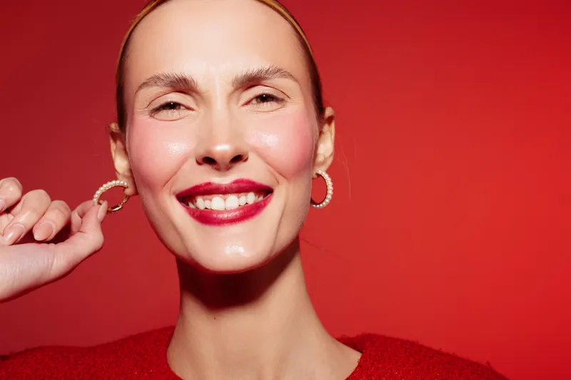 Smiling woman with red lipstick and hoop earrings poses against red background. Close-up portrait with smooth skin and joyful expression. Concept of beauty, fashion, and confidence.