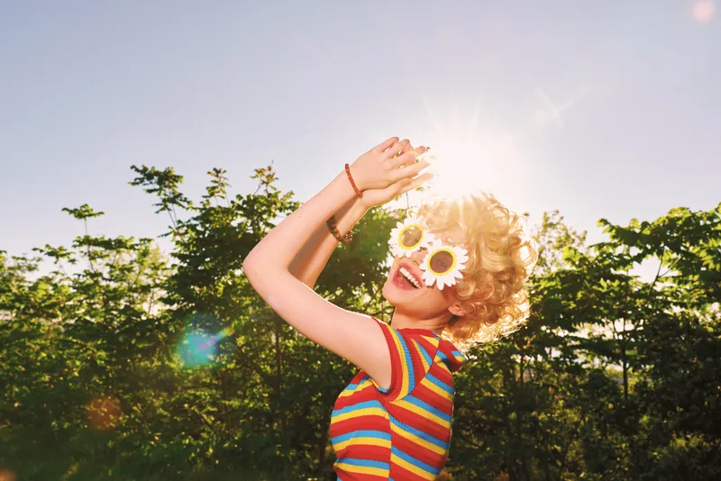 Joyful girl outdoors in a striped shirt and flower sunglasses, sunlit moment, playful pose, warm retro tones, lush greenery background, carefree mood, natural colors, soft breeze, candid laughter