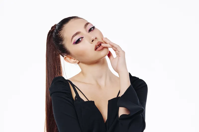 Young woman with dramatic pink eye makeup poses with fingers near lips wearing black off-shoulder top. High contrast beauty portrait on white background. Fashion and creative concept.
