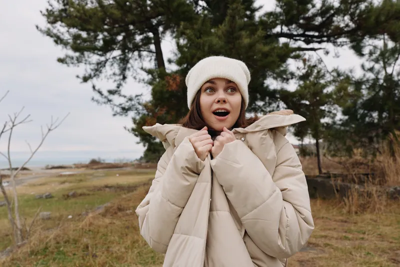 Woman in a beige puffer coat and knit beanie stands outdoors with a surprised expression, in a chilly field with trees and open landscape behind her.