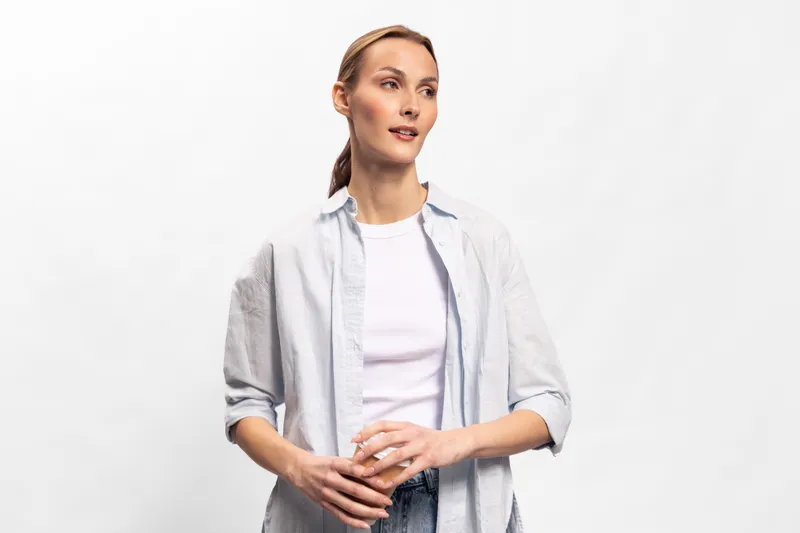 Young woman in casual white shirt stands against white background looking aside with slight smile. Natural light highlights her relaxed posture and thoughtful expression.